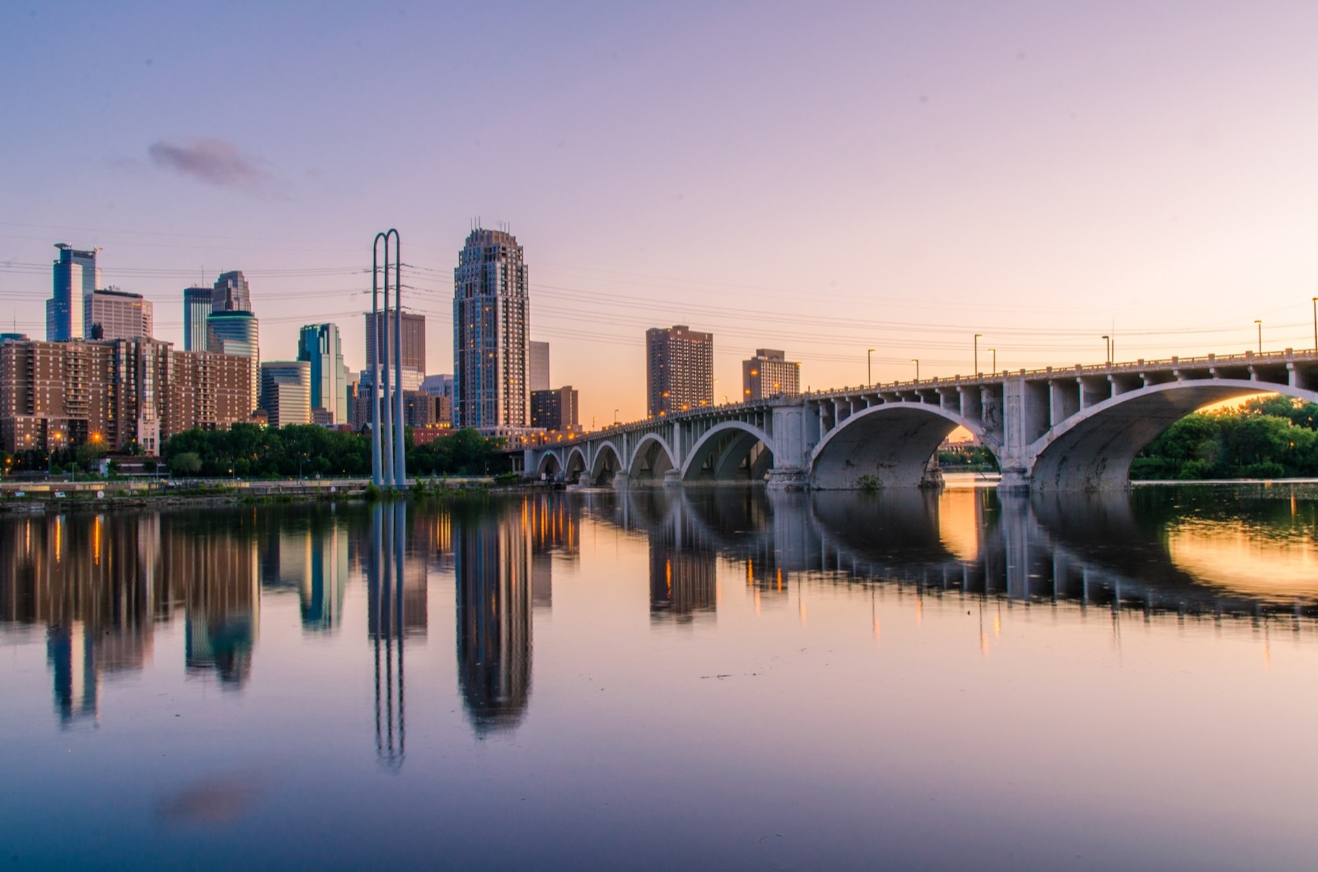 An arched bridge leading toward a city skyline, with water reflecting a lilac sky above; labor day weekend 