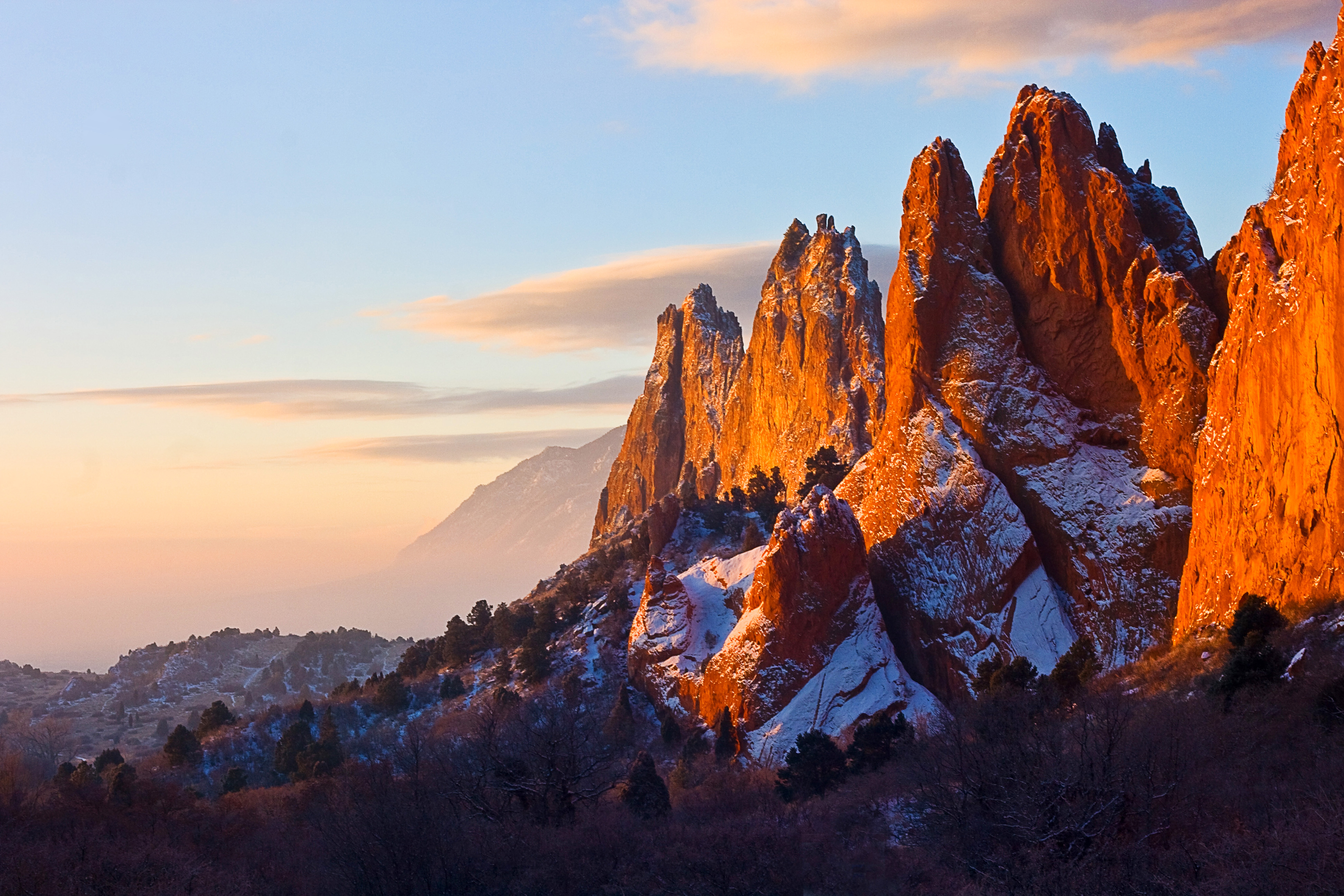 A dusting of snow covers several red rock pointed rocky peaks that rise above a hilly landscape