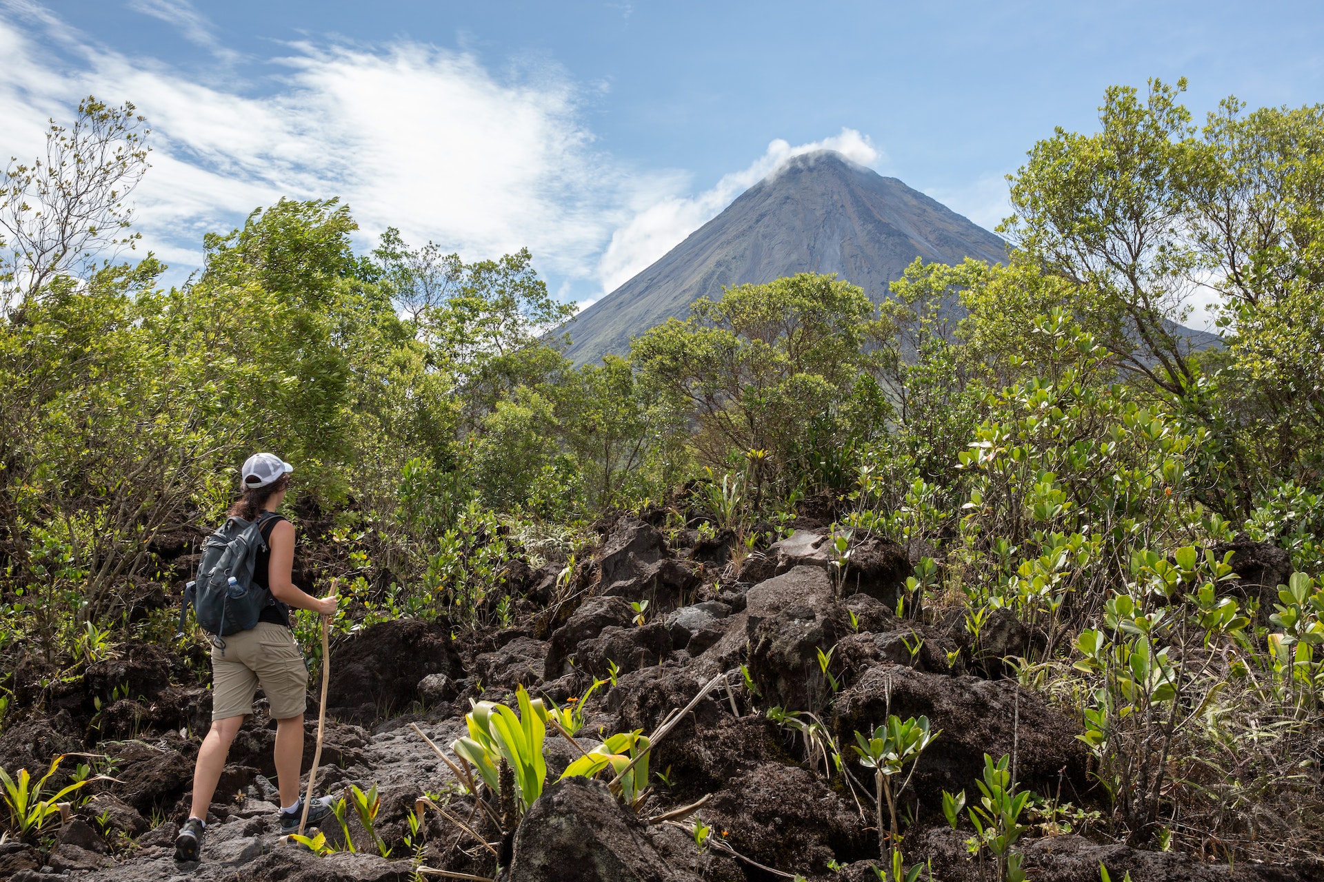 A hiker with a stick pauses on a trail to look at the huge volcano that dominates the jungle scenery