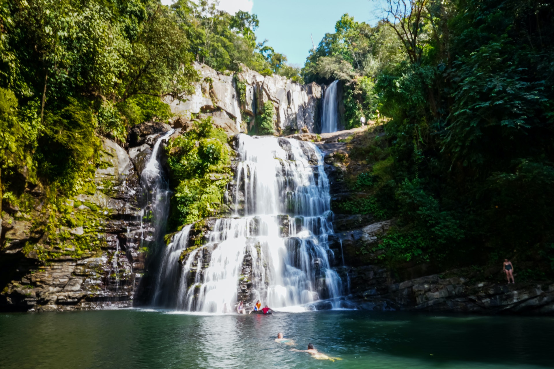 People swim and play in the water at the foot of a tiered watefall in a jungle